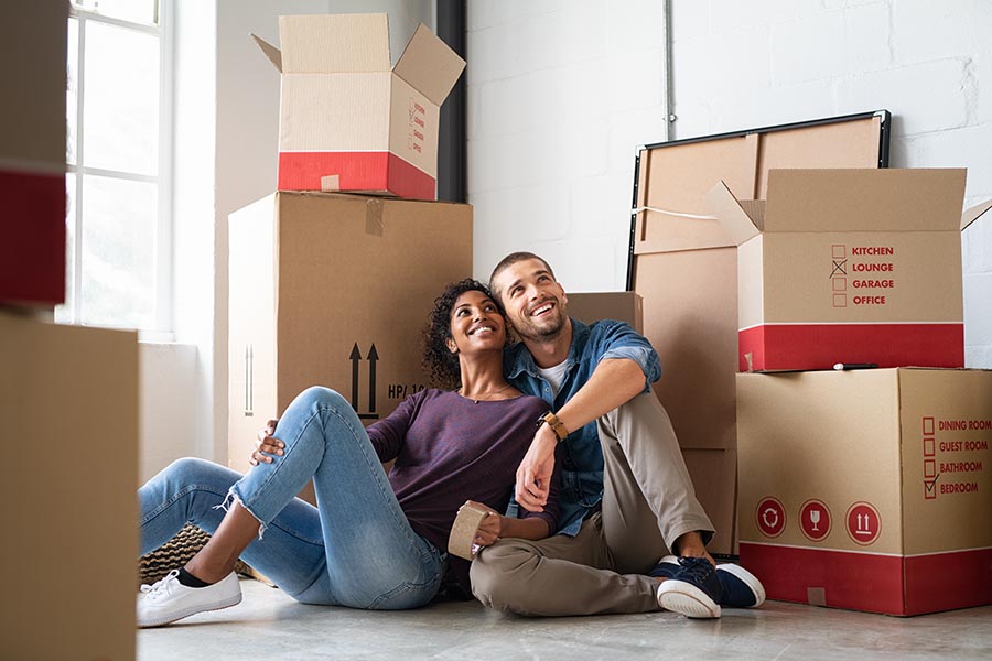 About Our Agency - Young Couple Sit Close Together Amongst Moving Boxes in Their New Home, Looking Proud